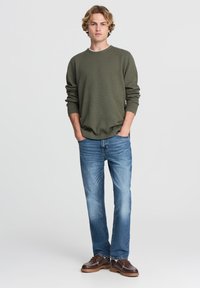 Zielono-oliwkowy, teksturowany sweter z długim rękawem; zestawiony z wygodnymi, jasnoniebieskimi dżinsami i brązowymi sznurowanymi butami z białym akcentem w szwie.