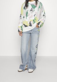 Barevný pletený svetr s abstraktními vzory v růžové, zelené a žluté barvě, doplněný světle modrými širokými džíny s motivy slunce a usměvavých obličejů.