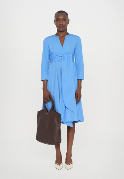 Abito blu avvolgente con maniche lunghe, una cintura in vita e una gonna a campana; abbinato a una borsa a tracolla in pelle marrone e scarpe bianche a punta.