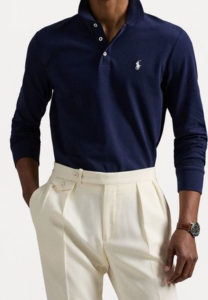 Homme portant un polo bleu marine à manches longues avec un logo blanc, un pantalon plissé crème avec un bouton latéral, et une montre-bracelet marron au bras gauche.