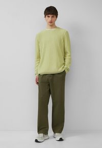 Lichtgroene geribde trui met een ronde hals, gecombineerd met donkerolijfgroene broek en grijze sneakers. Eenvoudig ontwerp, gestructureerde stof. Neutrale achtergrond.