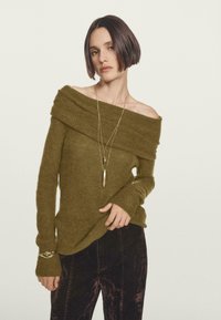 Frau mit schulterlangem, dunklem Haar, die einen olivgrünen, schulterfreien Pullover, lange goldene Halsketten, ein Armband und dunkel strukturierte Hosen vor einem weißen Hintergrund trägt.