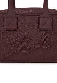 Sac à main en cuir bordeaux avec une finition texturée, des poignées supérieures doubles et un logo en relief. Forme rectangulaire avec des accents de couture.