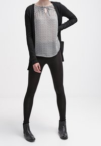Svart lång cardigan över en mönstrad vit blus med nyckelhålsutskärning, i kombination med svarta tight leggings och ankelstövlar.