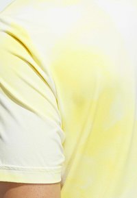 Krátký rukáv žlutého trička s texturovaným gradientním designem, který přechází od světlé žluté po bílou, s kulatým výstřihem.