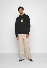 Sweat à capuche noir avec un logo graphique vert, associé à un pantalon cargo beige avec des poches. Le modèle pose sur un fond uni.