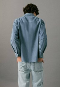 Ljusblå skjorta med knappar och långa ärmar, bakvy. Tyget är slätt med krage och synliga knappar vid muddarna. Bärs med ljusa jeans.