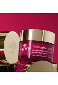 Pot van NUXE Merveillance Lift Firming Velvet Cream met een glazen roze lichaam en een gouden deksel, weergegeven tegen een heldere roze achtergrond.