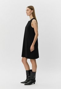 Zwarte mouwloze jurk met een ronde halslijn en decoratieve versieringen. Gecombineerd met glanzende zwarte enkellaarsjes met een krokodillenstructuur.