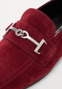 Punane sametist loafers, millel on hõbedase tooni metallist pandla ja aksessuaar, tekstureeritud pind ning suletud disain.