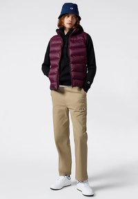 Bordowa pikowana kamizelka na czarnej bluzie z kapturem, beżowe spodnie, białe trampki i niebieski kapelusz typu bucket. Kamizelka ma pikowany wzór i wysoki kołnierz.