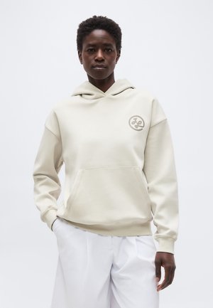 Osoba s krátkymi kučeravými vlasmi, oblečená v svetlo béžovej kapucni s predným vreckom a malým kruhovým logom na hrudi, v kombinácii s bielymi nohavicami.