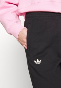 Osoba oblečená v černých teplákách Adidas s bílým logem a růžové mikině, ruka opřená v kapse tepláků.