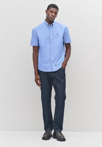 Chemise bleu clair à boutons avec manches courtes, associée à un pantalon en denim foncé et des chaussures noires, ornée d'un petit logo brodé sur la chemise.