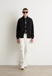 Jachetă zip neagră cu două buzunare pe piept, purtată peste o cămașă cu carouri, asociată cu pantaloni albi și adidași albi. Modelul poartă ochelari de soare.