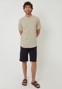 Camisa corta de manga con cuello mao en color beige claro y botones al frente, combinada con pantalones cortos oscuros y sandalias marrones sobre un fondo liso.