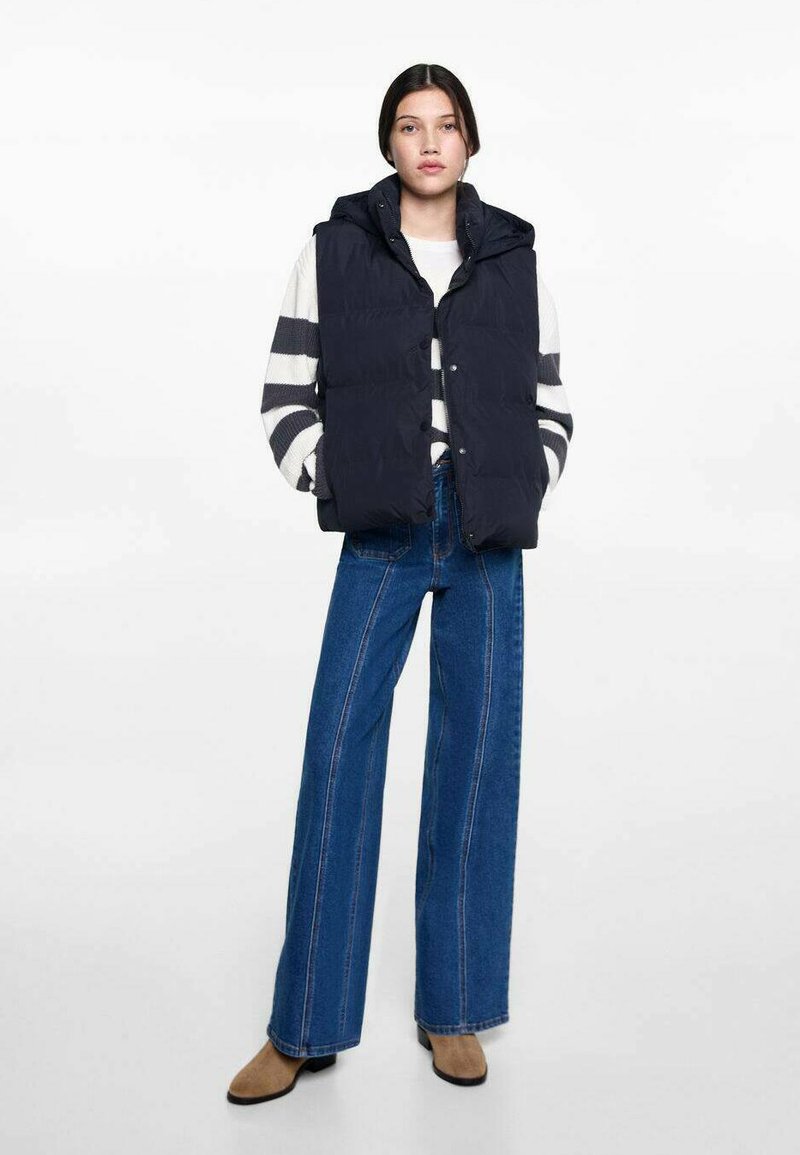 Marinblå puffväst med huva, knappstängning, bärs över en randig tröja. Kombineras med vida blå jeans och bruna ankelstövlar.