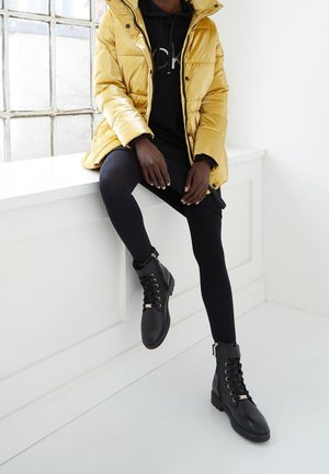 Personne portant une doudoune jaune, un sweat à capuche noir, des leggings noirs et des bottes noires à lacets, assise sur un rebord de fenêtre blanc dans une pièce lumineuse.