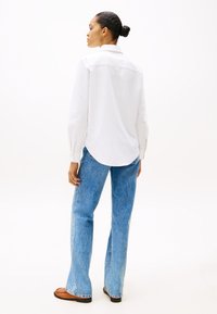 Vista trasera de una persona que lleva una camisa blanca de manga larga, jeans acampanados de color azul claro y zapatos planos marrones sobre un fondo blanco.