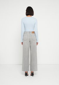 Ανοιχτό μπλε μακρυμάνικο τοπ συνδυασμένο με γκρί τζιν ψηλόμεσο, με φαρδιά μπατζάκια. Διαθέτει ελάχιστες ραφές και ομοιογενή υφή. Ορατά μαύρα τακούνια.