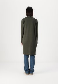 Lang, slim-fit coat in olijfgroen, gemaakt van zachte stof. Heeft lange mouwen en valt tot halverwege de dij, gecombineerd met spijkerbroek en lichte schoenen.