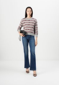 Maglione grigio con motivo "JOOP! JEANS" in nero e rosso, abbinato a jeans in denim blu a zampa d'elefante e sandali neri con tacco alto. Tiene in mano una piccola borsa nera.
