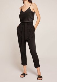 Mujer vistiendo un top de satén negro, pantalones cortos de cintura alta con textura y cinturón, y sandalias negras de tiras abiertas, de pie con las manos en los bolsillos.
