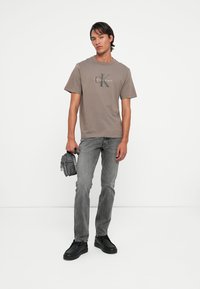 Gray cotton t-shirt with a logo print, paired with gray jeans and black sneakers. The model holds a small black bag. Simple design, casual fit.