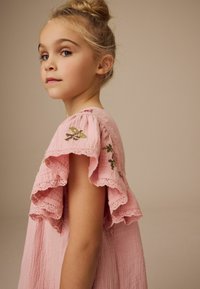 Rosa Kleid mit Spitzenbesatz, Rüschensleeven und floralen Stickdetails. Weiche Textur und lockere Passform, die gerade unter die Schultern fällt.