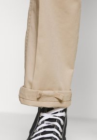 Béžové zrolované kalhoty z bavlněného kepru, s hladkou texturou a přizpůsobeným střihem, spárované s černými vysokými teniskami s bílými tkaničkami.