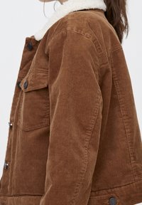 Donna che indossa una giacca in corduroy marrone con colletto in pile bianco e bottoni neri, mostrata di lato all'altezza del torso medio.