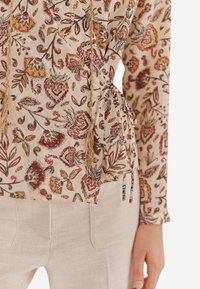 Bluse med blomsterprint i lys beige stof, der har indviklede røde og gule designs, sidesnøringer og strukturerede overflader.