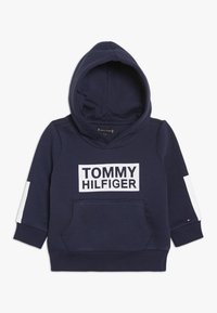 Sudadera con capucha azul marino con un gran logo blanco "TOMMY HILFIGER", bolsillo canguro frontal y detalles blancos en las mangas. Material de algodón.