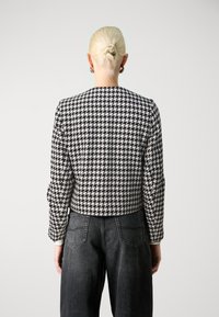 Svart og hvitt jakke med hundetannmønster, avrundet halslinje, lange ermer og kort design, kombinert med mørke denimbukser.
