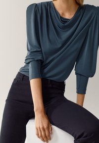 Blusa drappeggiata teal con maniche lunghe, caratterizzata da spalle plissettate e una texture morbida, abbinata a pantaloni scuri aderenti.