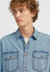 Light blue denim shirt with a button-up front, two chest pockets, brown snap buttons, and a classic collar, worn over a white shirt.