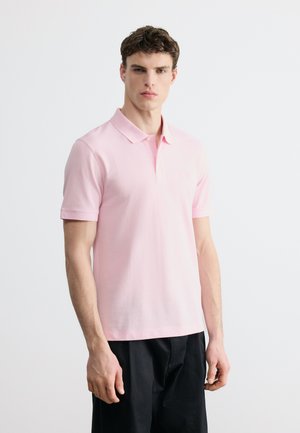 Mladý muž s krátkými kudrnatými vlasy, oblečený v světle růžové krátkosrsté polo košili a černých kalhotách, stojící před jednobarevným bílým pozadím.