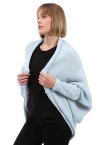 Hellblau gestrickter Schal mit geripptem Muster, offenes Design und lockere Passform, über einem schwarzen Oberteil getragen, präsentiert von einem Modell.