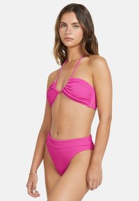 Horúco ružový bikiny set obsahujúci textúrovaný bandeau top s uzlom a vysokými nohavičkami. Hladká, elastická tkanina, minimálne kovové prvky.