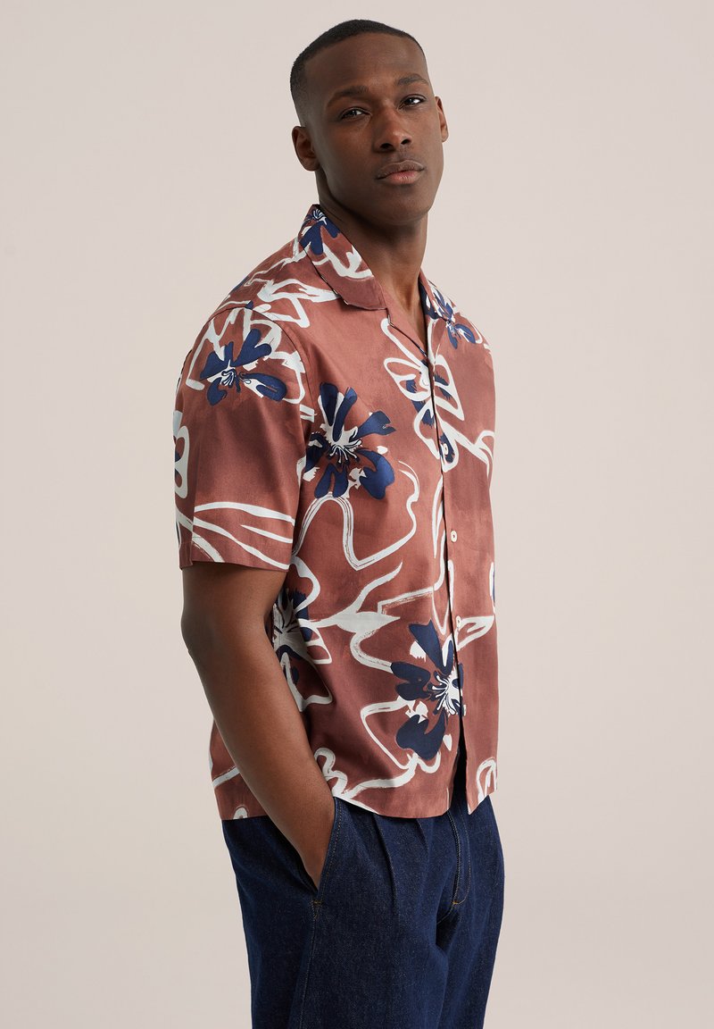 Homme portant une chemise à manches courtes boutonnée avec de grands motifs floraux bleu marine et blanc, associée à un pantalon bleu foncé, mains dans les poches.