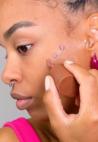 Kosmetisk stift i brun påført kinnet, med tre rosa prikker på huden. Personen har septumpiercing og har på seg rosa antrekk.