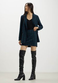 Stivali neri in pelle sopra il ginocchio, con punta quadrata e tacco a blocco; abbinati a un blazer a quadri verde scuro e una gonna coordinata sopra un top nero.