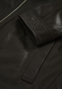 Primo piano della manica e della tasca di una giacca di pelle marrone scuro con texture della grana visibile e dettagli di cucitura.