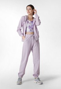 Felpa viola chiaro con zip frontale e tasche, abbinata a pantaloni tuta con cordino coordinati. Modella che indossa un top corto floreale e sneakers.