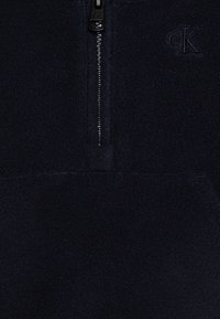 Suéter de fleece de media cremallera en azul marino oscuro con tejido texturizado y logo bordado. Presenta una cremallera suave y un bolsillo canguro.