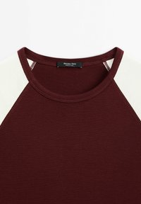 Burgunder T-shirt med cremefarvede raglanærmer, rund halsudskæring og et vævet mærke i midten af kraven. Blød, glat tekstur.
