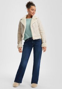 Veste en shearling beige clair avec un col, associée à un t-shirt côtelé bleu-vert et un jean évasé bleu foncé. Chaussons beiges en peluche.