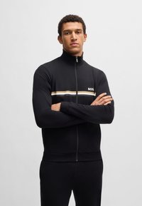 Schwarze durchgängige Sportjacke mit hohem Kragen, weißen und goldenen horizontalen Streifen sowie einem Logo auf der linken Brust. Glatte Textur.