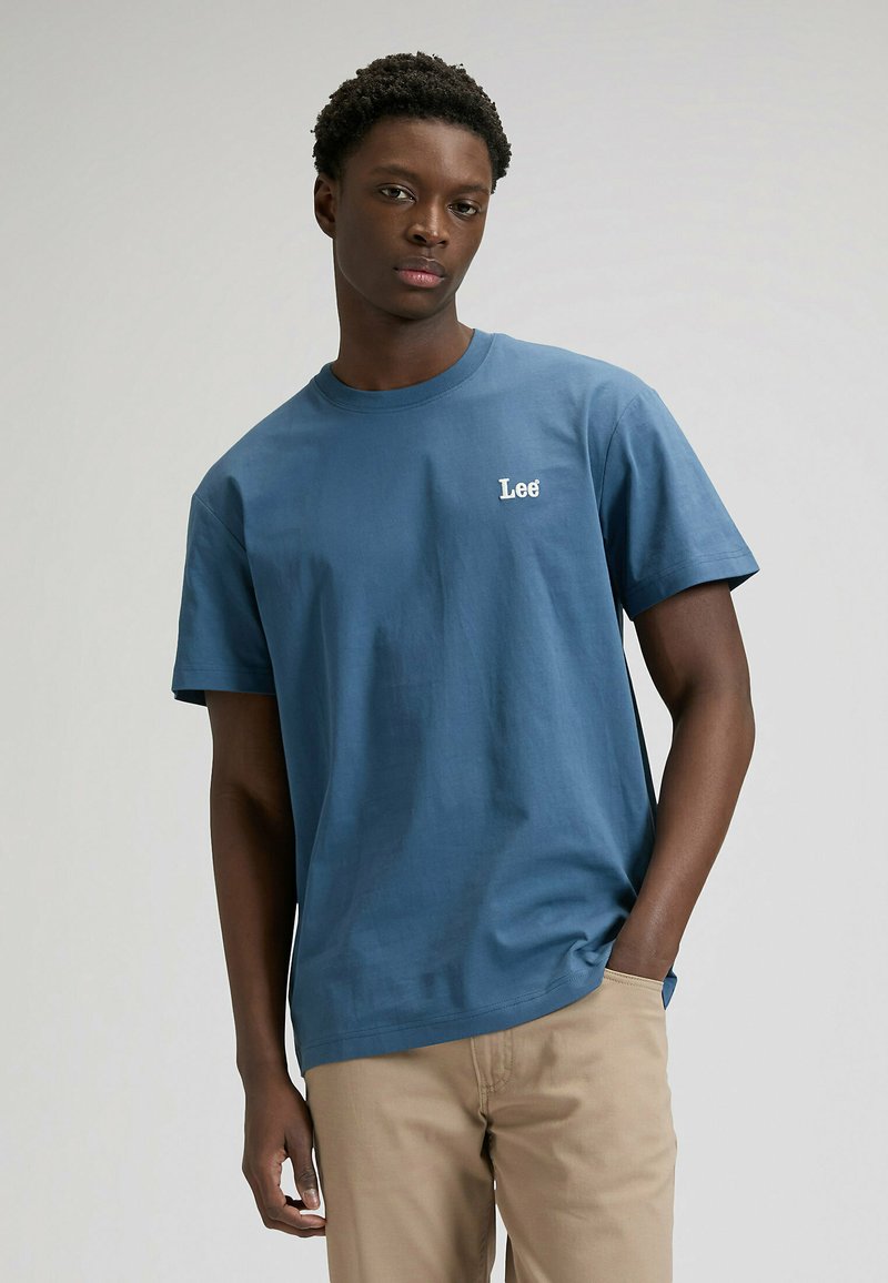 T-shirt blu in cotone a maniche corte con collo rotondo e logo "Lee" bianco sul petto, abbinata a pantaloni beige. Texture liscia, vestibilità casual.