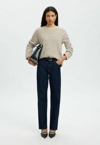 Femme se tenant debout avec des cheveux bouclés, portant un pull beige côtelé, un jean bleu foncé, des talons noirs, tenant un sac à main noir sur fond uni.